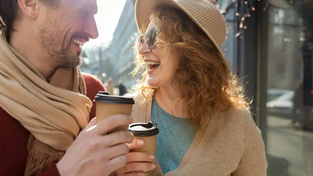 A couple laughing while holding coffee cups outdoors.