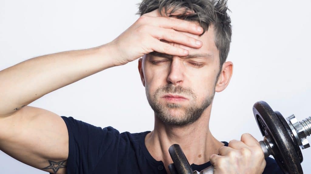 A lightly bearded man with closed eyes holding his forehead wiht one hand and a dumbbell in the other.