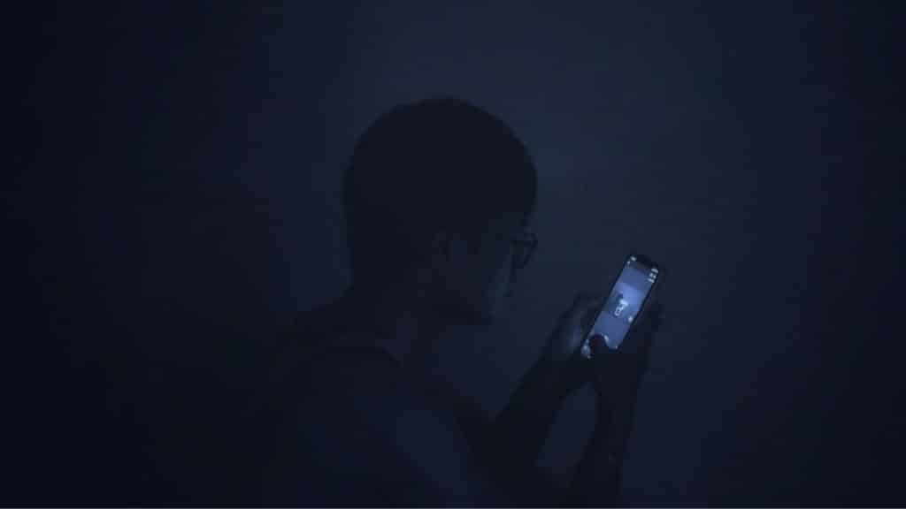 A person in silhouette sits in a dark room looking at a glowing smartphone screen.