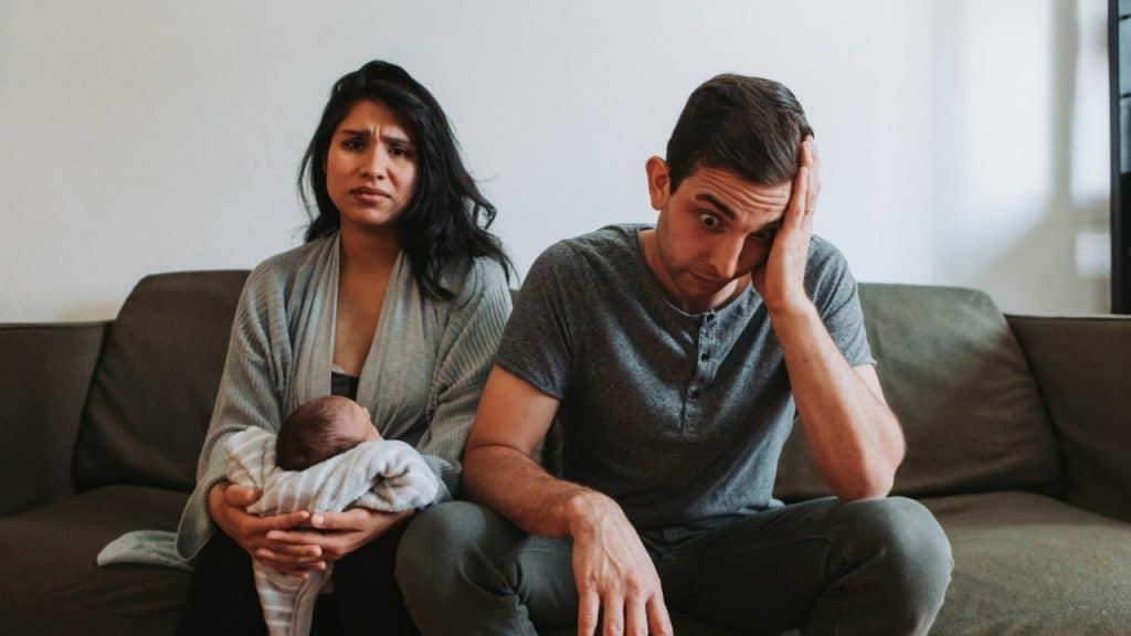 A woman holding an infant sits beside a man with his hand on his head.