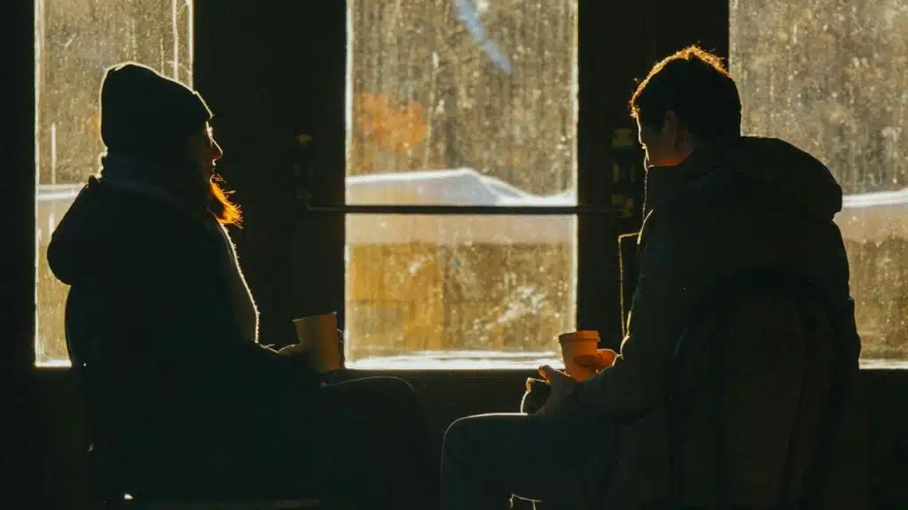Silhouettes of two people holding mugs sit facing each other in front of a window.