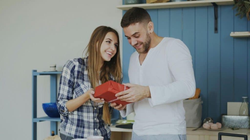 A woman smiles as she gives a red wrapped gift to a man indoors.