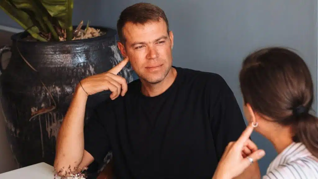 A man in a black shirt looks toward a woman while pointing at his ear.