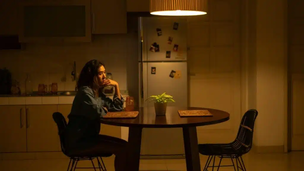 A woman sits alone at a round wooden table in a dimly lit kitchen at night.