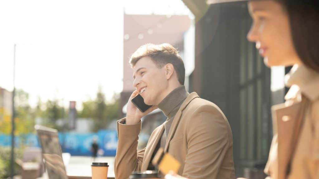 A man in a tan jacket smiles while talking on a smartphone at an outdoor table.