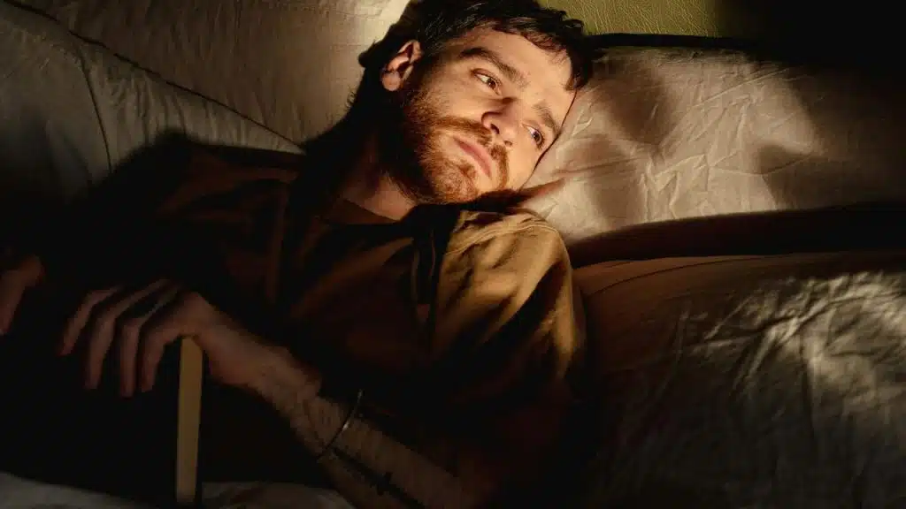 A bearded person lying awake in bed, softly lit and gazing off to the side.