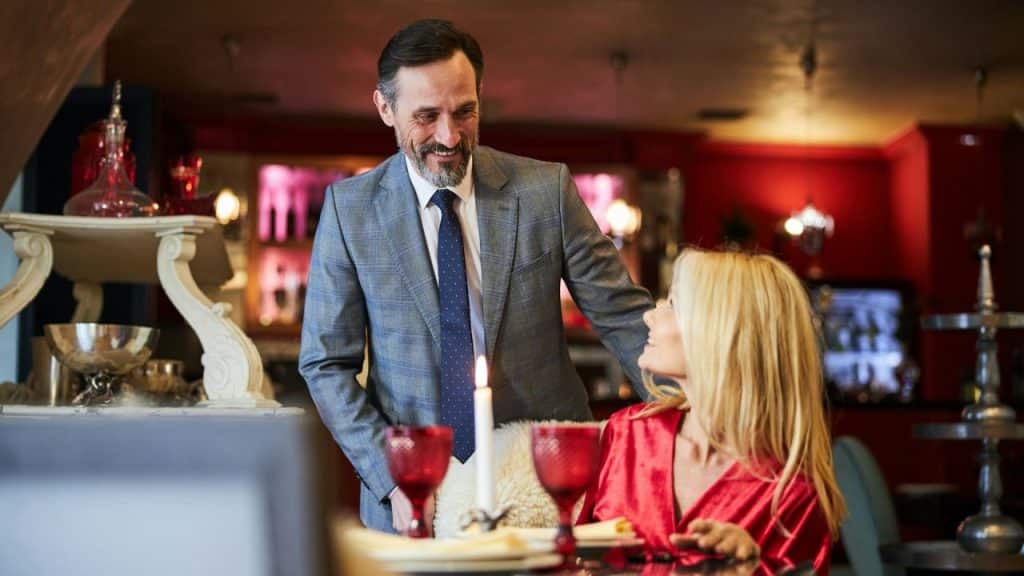 A man in a suit smiles down at a woman seated at a restaurant table.
