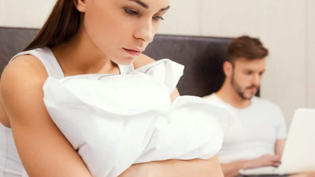 A woman clutches a white pillow while a man works on a laptop behind her.