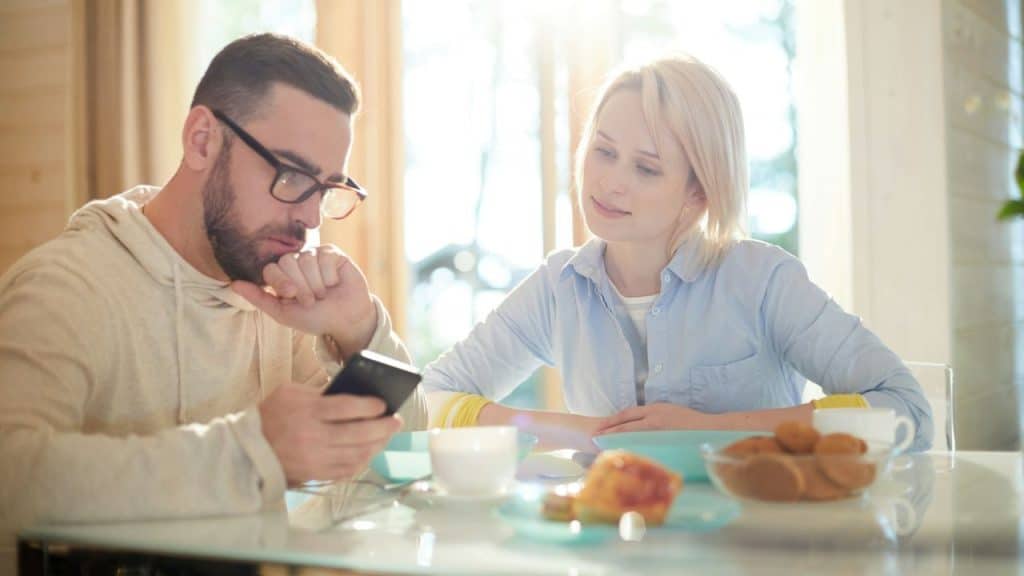 A woman looks at a man who is focused on his phone while sitting at a table.