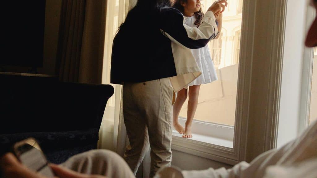 A parent lifting their young daughter in the air near a sunny window in their home.