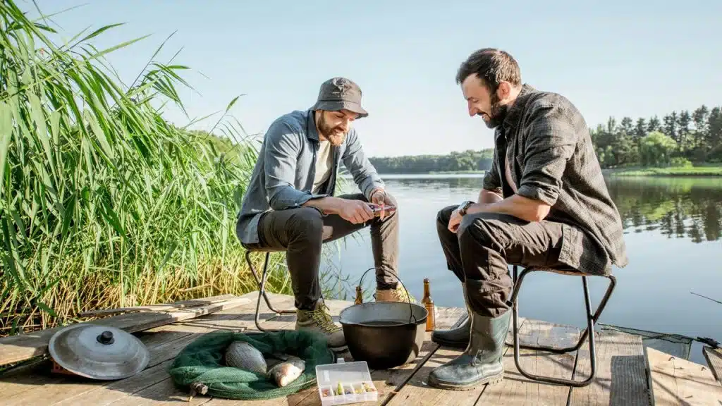 A pair of men sitting by a lakeside dock while fishing and chatting.