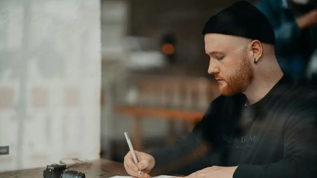 A man in a beanie writing at a table with a camera nearby.
