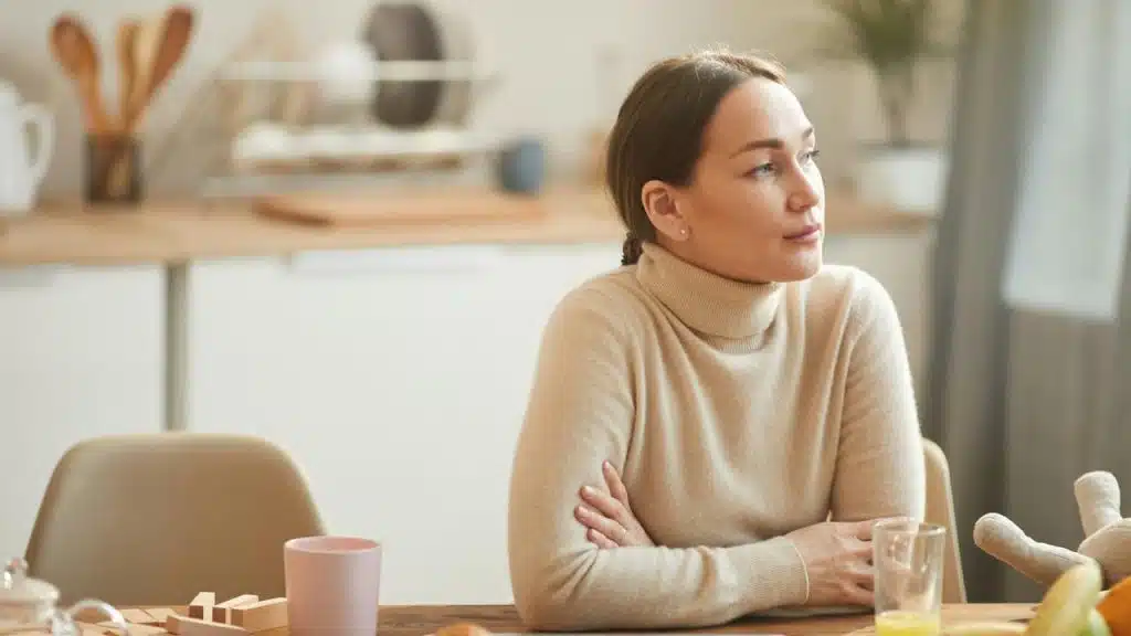 A woman sitting thoughtfully at a kitchen table in a bright home.
