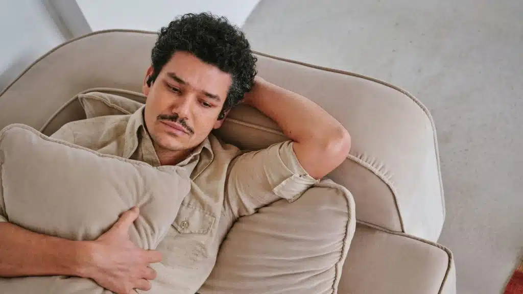 A man with curly dark hair and a mustache reclines on a tan sofa holding a pillow.