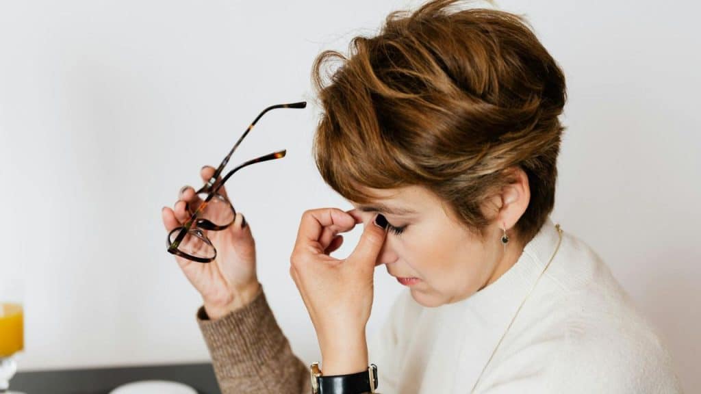 A woman with short brown hair holds glasses while pinching the bridge of her closed eyes.