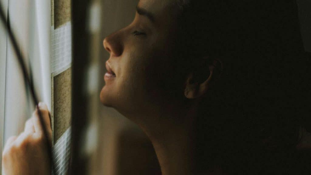 A person in close-up touching window blinds with eyes closed.