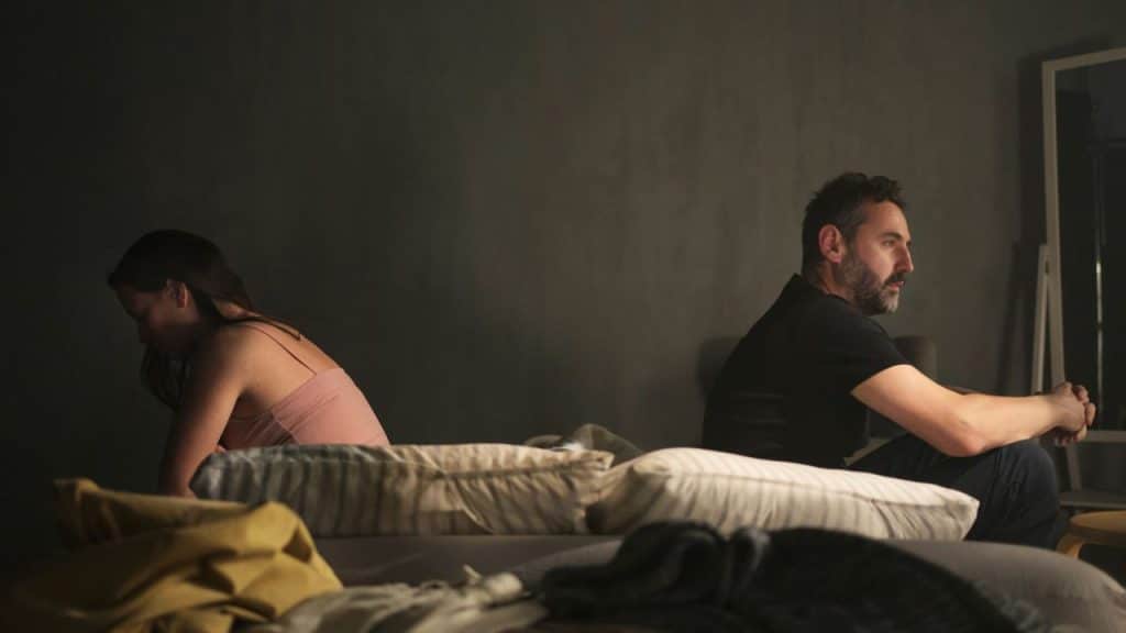 A man and woman sit on a bed facing away from each other in a dark room.