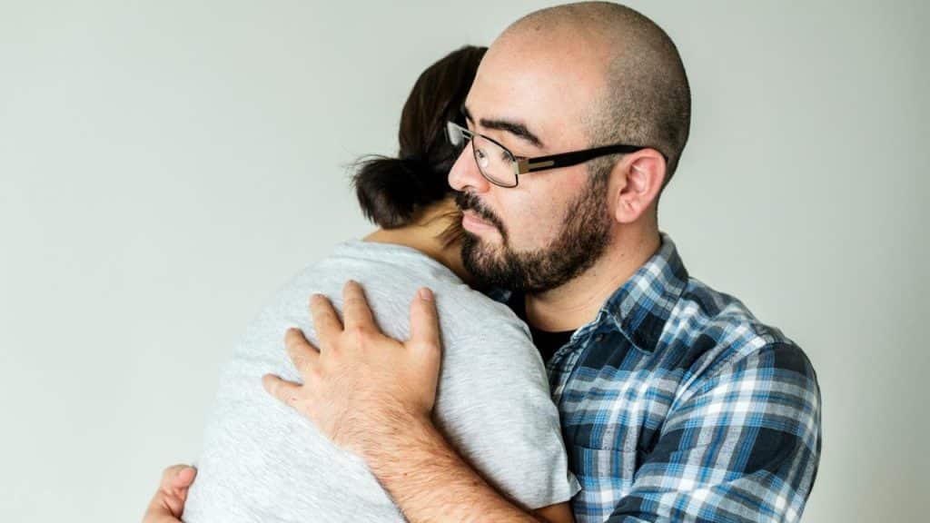 A man with a beard and glasses embraces a person whose back is to the camera.
