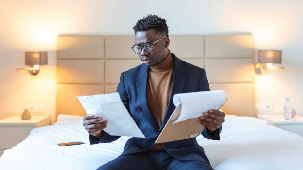 A man in a suit and glasses sits on a bed reviewing papers on a clipboard.
