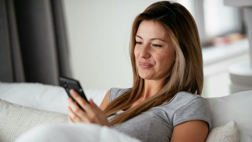 A woman with light brown hair sits on a white couch while looking at her smartphone.