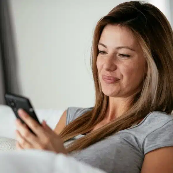 A woman with light brown hair sits on a white couch while looking at her smartphone.