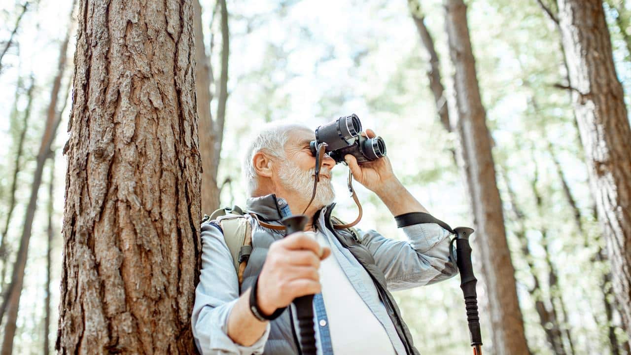 An elderly hiker leaning against a tree looking through binoculars.