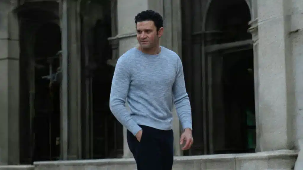 A man in a grey sweater walks outdoors with one hand in his pocket, looking aside.