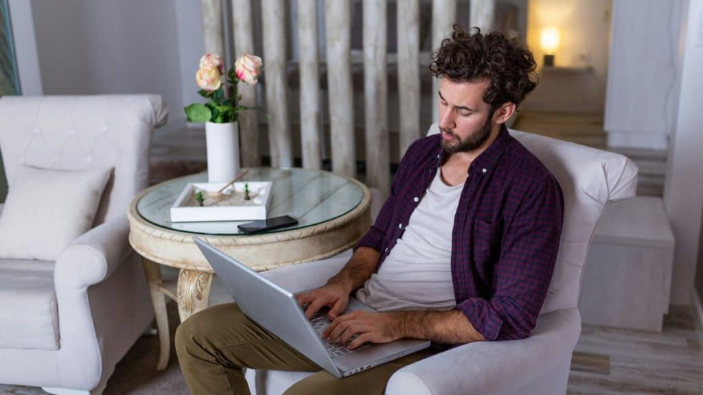 A man with curly hair sits in a white armchair using a laptop near a table.