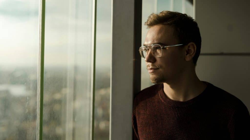 A man wearing glasses looks out of a large window toward a blurry city view.