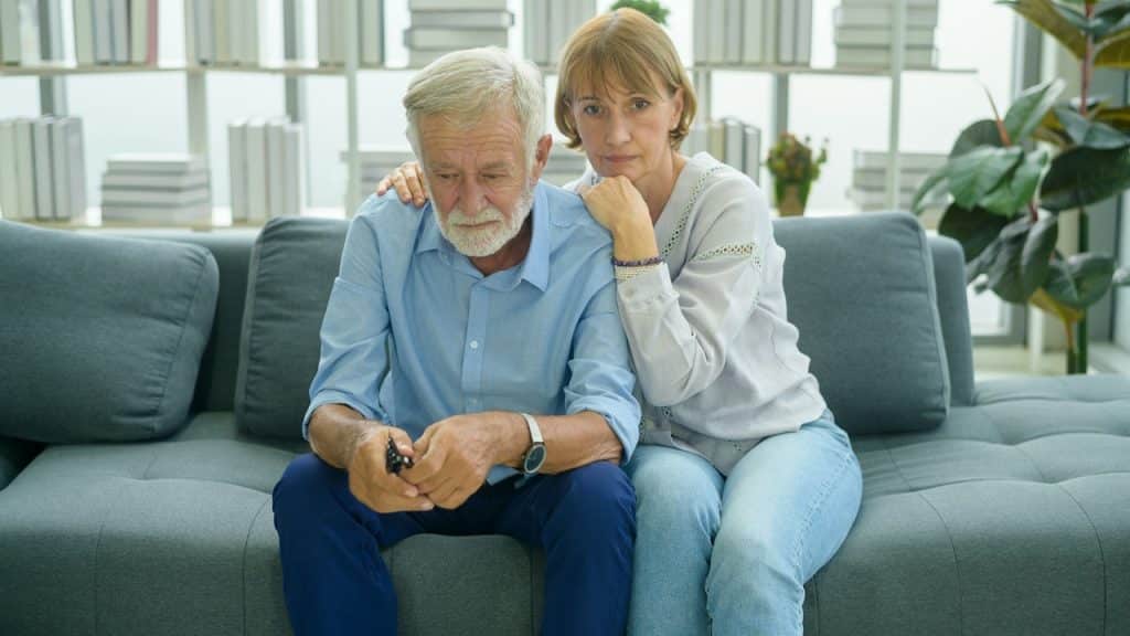 An elderly couple sitting on a couch as the woman holds her husband close.