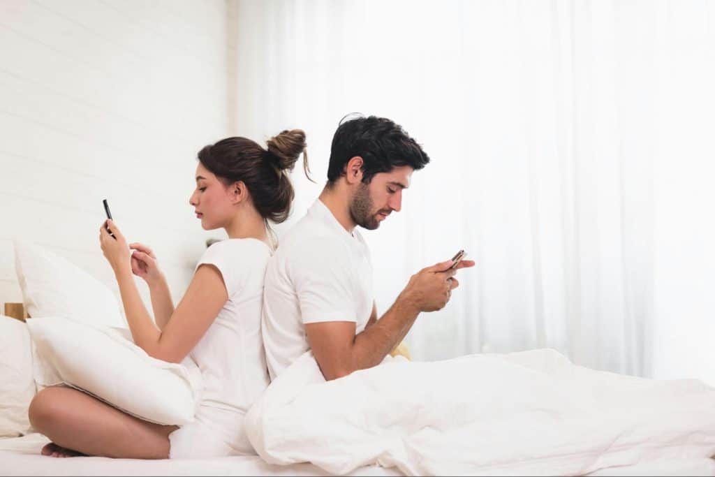 A couple sitting at the bed back to back and using their phones