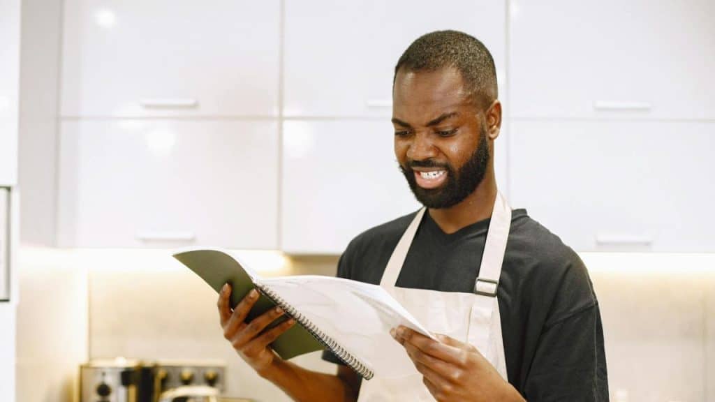A man wearing an apron looks down at an open book in a bright white kitchen.