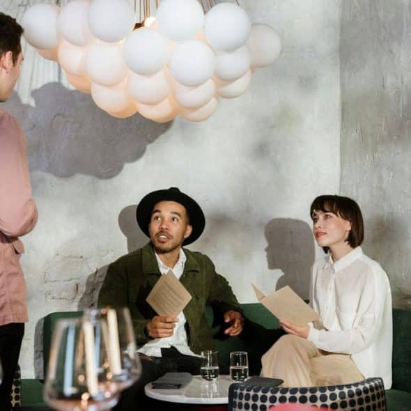 A man in a pink shirt stands before a seated couple holding menus under globe lights.