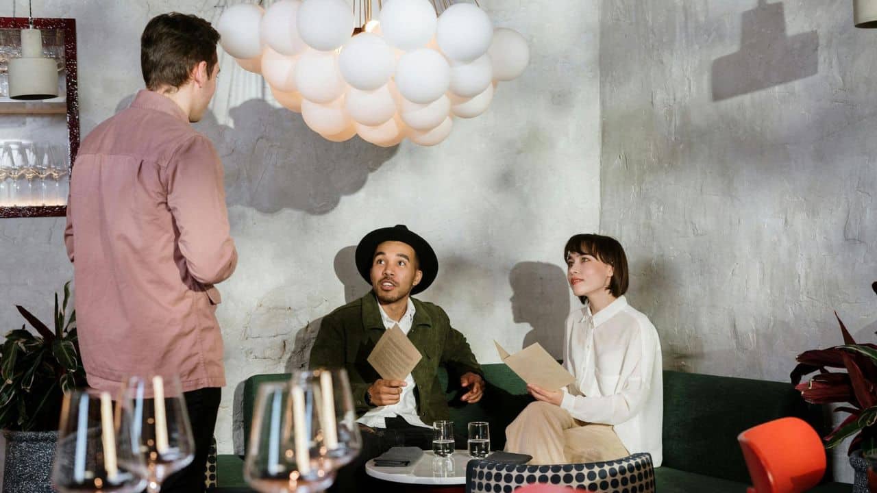 A man in a pink shirt stands before a seated couple holding menus under globe lights.