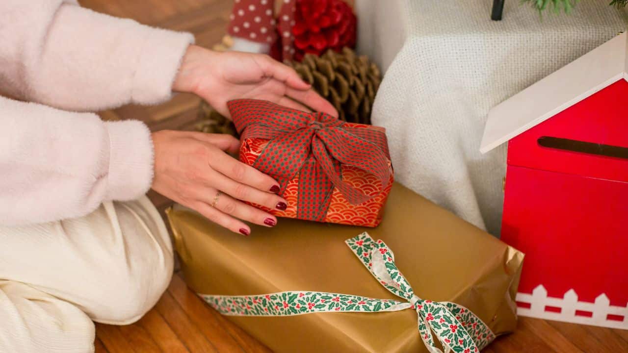 Hands place a small red wrapped gift on top of a larger gold wrapped gift box.