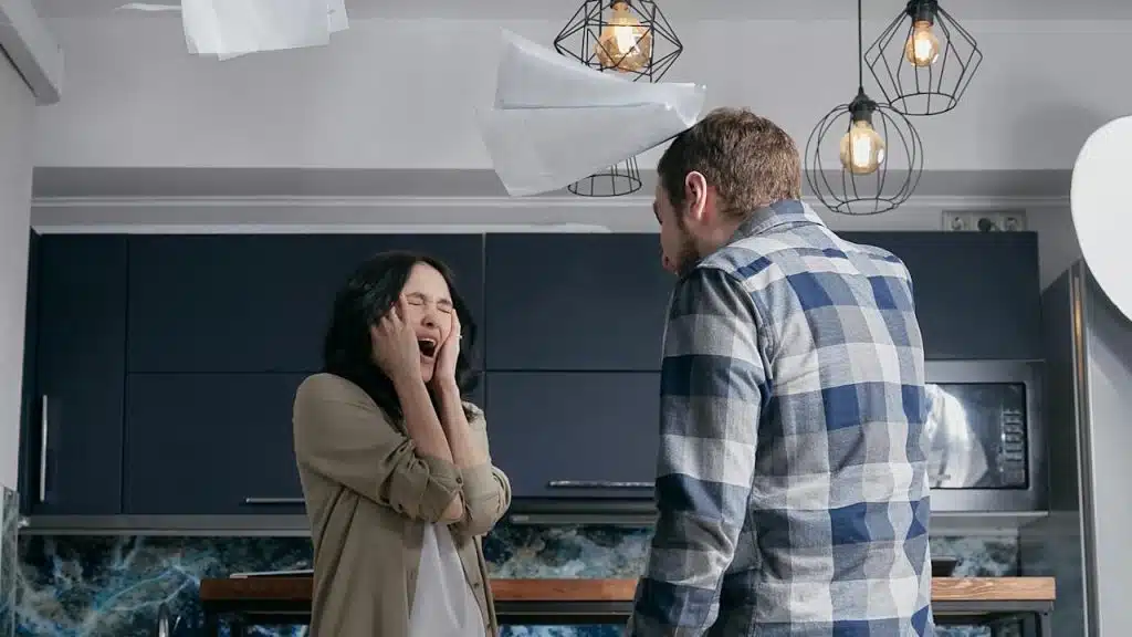 A woman screaming and holding her face in her hands while a man stands in front of her in a kitchen.
