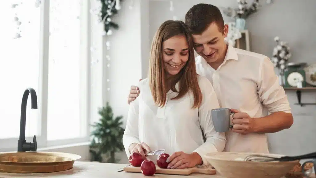 A couple smiling while preparing food together in a kitchen.