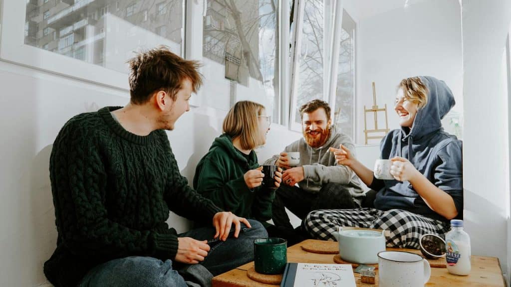 A group of friends laughing and drinking coffee together by a bright window.