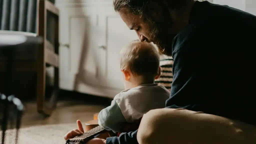 A father sitting with a young child while playing a small guitar indoors.