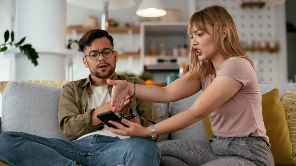 A woman gestures toward a smartphone while speaking to a man sitting on a sofa.