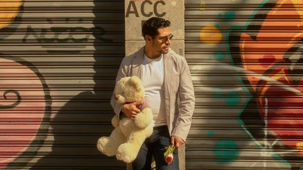 A man in a blazer holds a large teddy bear and a single pink rose.