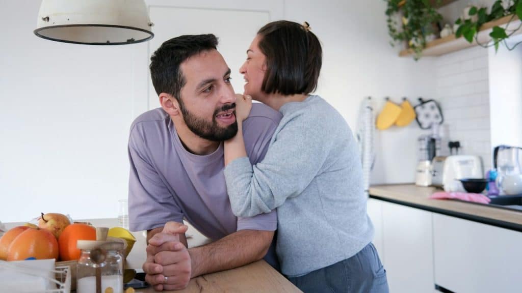 A woman hugs a man from behind in a kitchen while he looks toward the camera.