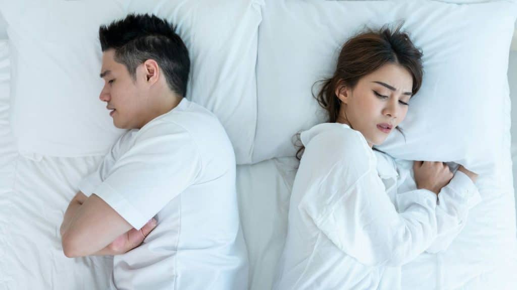 An Asian man and woman lie back-to-back on a white bed with eyes closed.