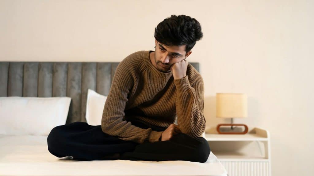 A man in a brown sweater sits cross-legged on a bed with his head on his hand.