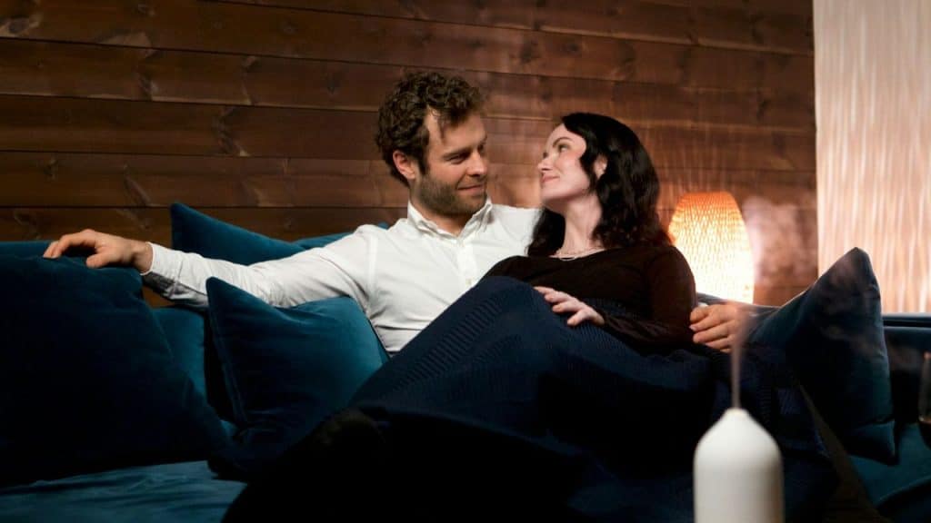 A man and woman sit closely together on a blue sofa, looking at each other.