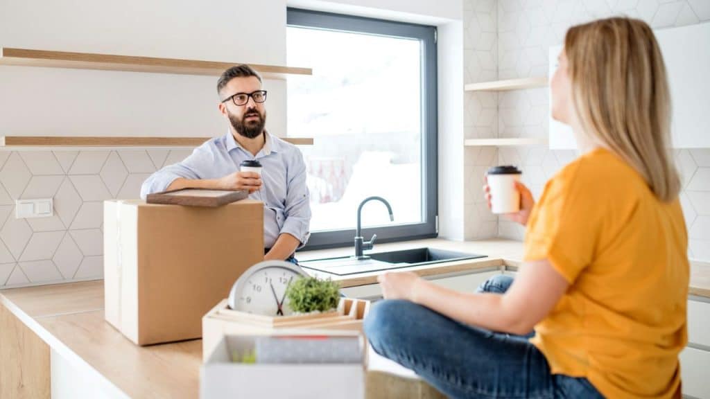 A man and woman sit in a kitchen with cardboard boxes and coffee cups.