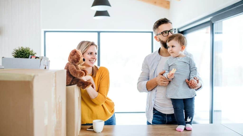 A family with a baby standing by moving boxes in a bright home.