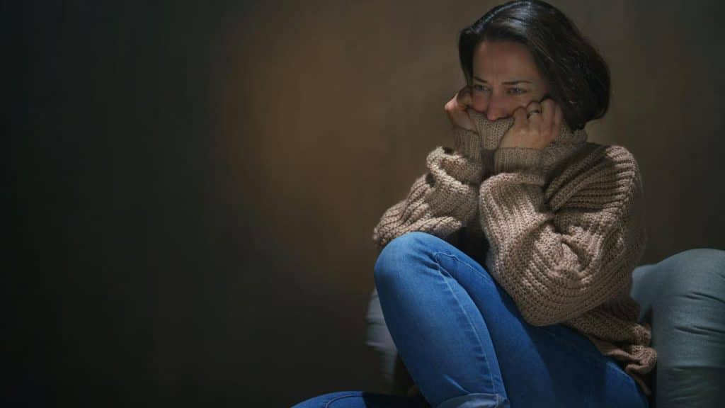 A woman sitting curled up in a sweater, holding her sleeves to her face and looking distressed.