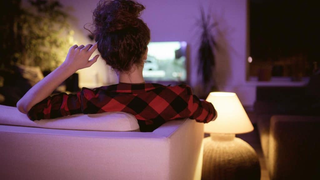 A woman watching TV while sitting on a couch on a subtly lit living room.