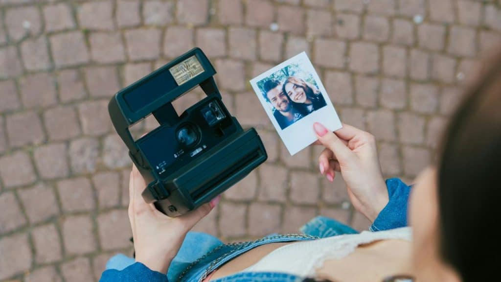 A person holds an instant camera and a small photograph of a smiling couple outdoors.